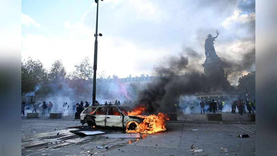 Veículo incendiado em praça na França durante protestos, com fumaça intensa e manifestantes ao fundo